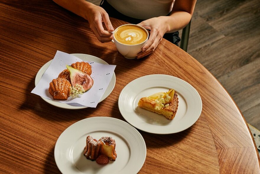 Bird's-eye of hand holding coffee cup, three plates of pastries on wooden table.