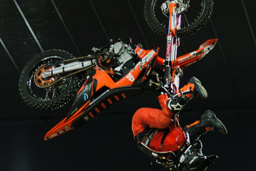A back flipping motocross bike doing a mid-air stunt, with the driver upside down but still holding the handlebars.