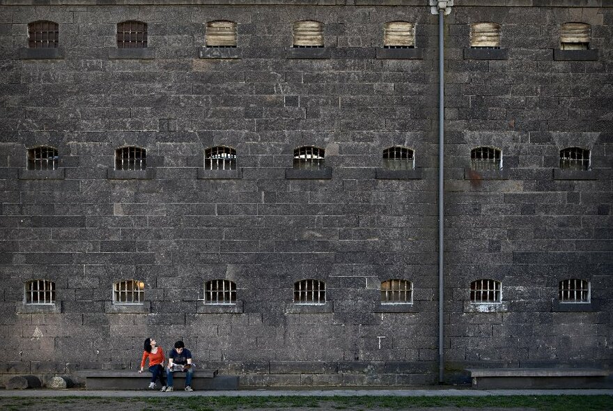 Two people sitting on a bench outside a gaol with barred cell windows. 