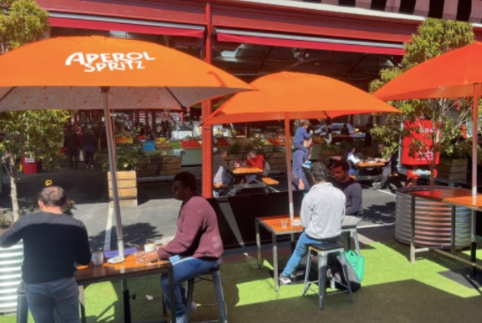 Diners seated at outdoor tables under orange umbrellas outside busy cafe.
