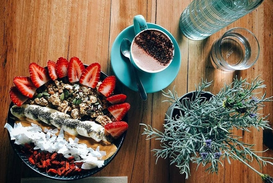 Granola with row of banana slices, strip of coconut and splayed strawberry pieces at edge of pool, cappuccino in a blue cup to the right, floral sprig to the bottom right. 