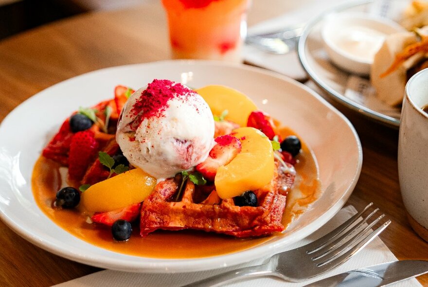 A white dish of waffles, poached fruit and ice-cream on a dining table in a cafe. 
