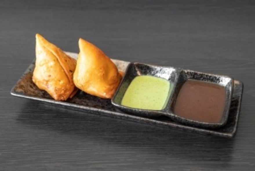 Two pakora next to dipping sauce on small, black rectangular platter.