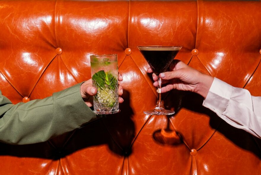 Two different hands, each holding cocktails, bringing the glasses close together in a celebratory fashion as if about to clink edges, against the backdrop of a rust-coloured leather banquette backrest.
