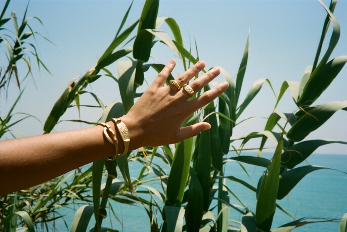 A hand and arm with rings and bracelets photographed against green foliage, blue sea and sky.