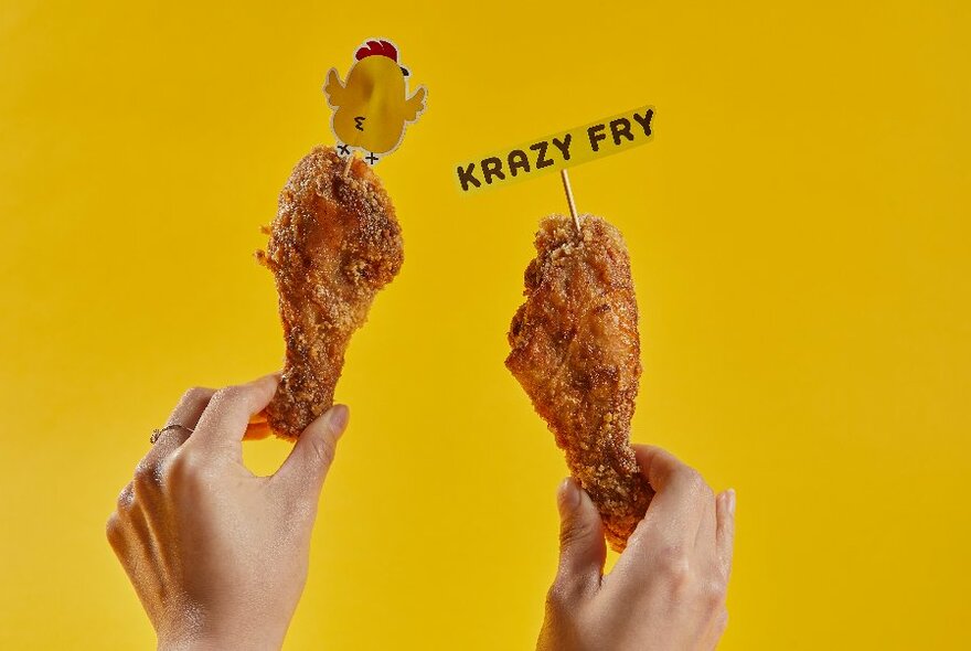 Two hands holding crispy fried chicken drumsticks, one with a little, paper chick stuck on top, the other with yellow piece of paper on a toothpick with words 'KRAZY FRY'.