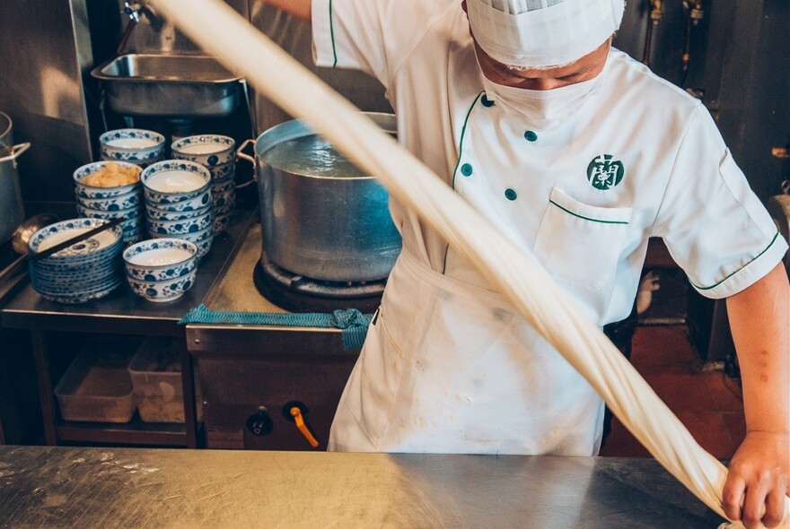 Chef stretching long piece of noodle dough above a stainless-steel bench, with blue floral bowls in background.