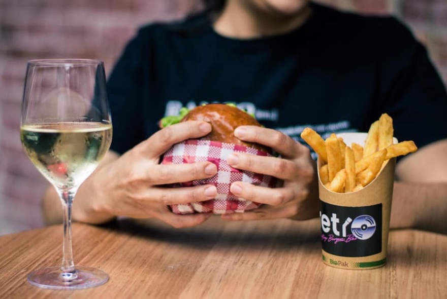 Glass of white wine, hands holding a burger in front of black T-shirt clad body, chips in a bucket with Retro name and logo.