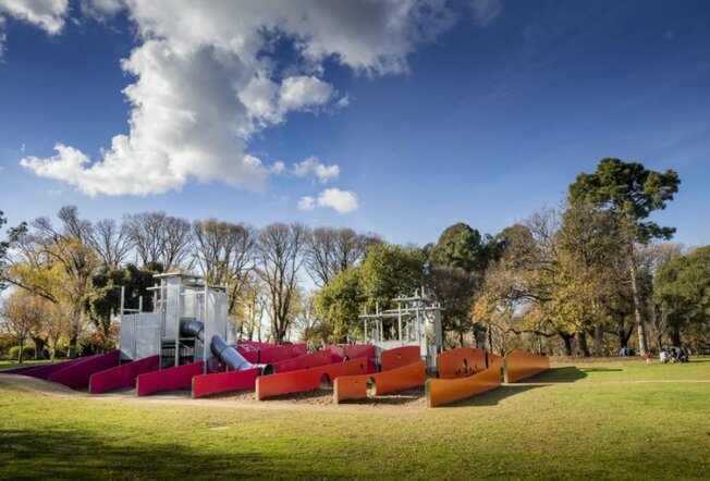 A playground with red and orange walls forming a maze in a green park. 