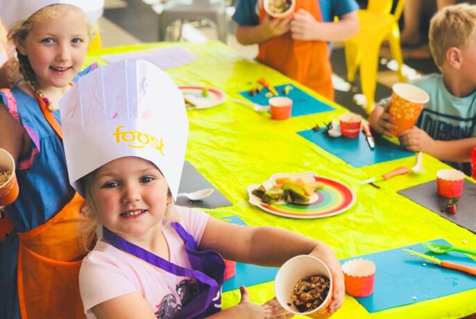 Preschool aged children wearing paper chef's hats and participating in a food/cooking class around a table.