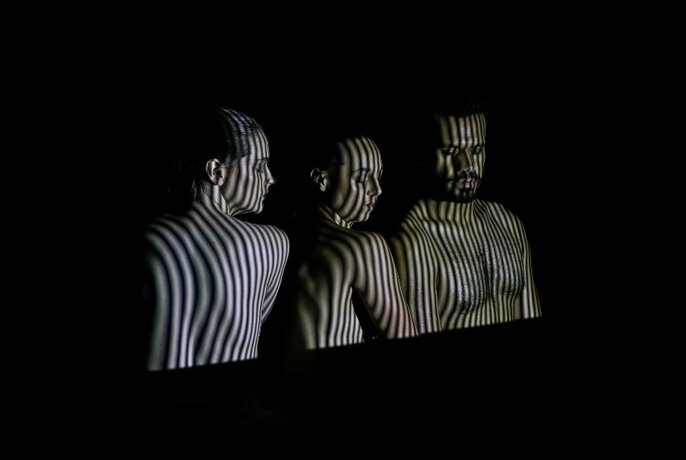 Three performers with striped lighting and shadows superimposed across their naked torsos.