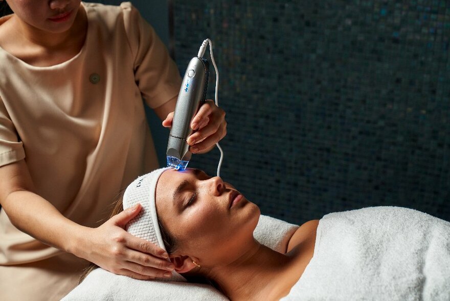 Therapist placing a beauty massage tool on the forehead of a customer lying on a table, head and body wrapped in white towels.