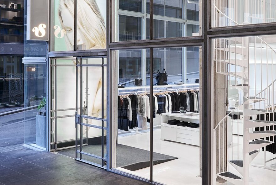 The glass entrance to a fashion store, a glimpse of black and white clothes hanging on racks, and a white spiral staircase to the right.