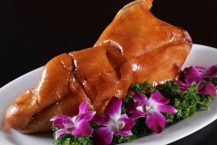 Glazed whole Peking duck on a white platter with purple flowers and parsley.