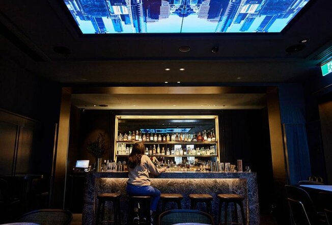 A woman sitting in a dark hotel bar with a projection on the roof.