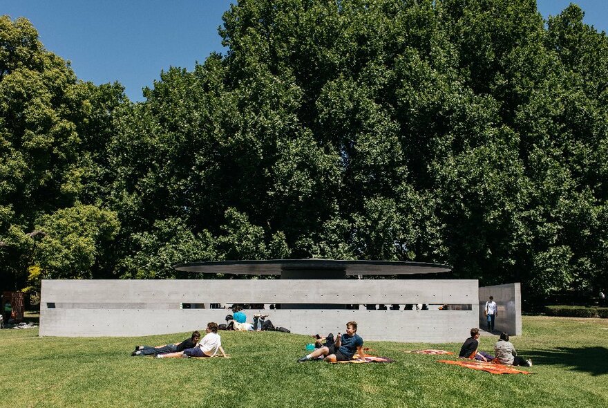 Melbourne's low concrete MPavilion in the Queen Victoria Gardens surrounded by trees and green lawn, with people lazing on the grass in front.
