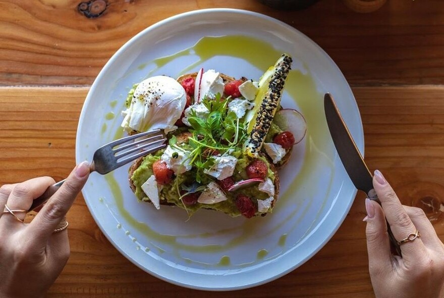 Hands holding cutlery above a plate with salad, poached egg and toast drizzled with olive oil.