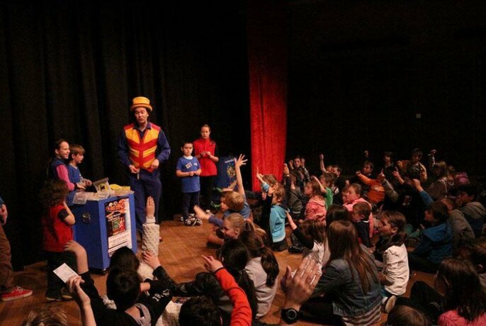 Performer standing on stage with several children with an audience of mostly children watching.