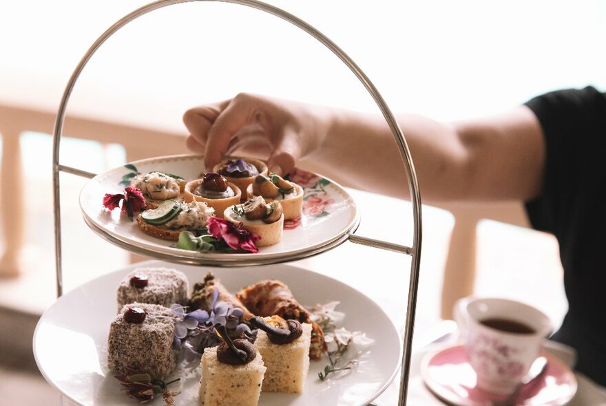 A hand hovering near a tiered plate of assorted sweet and savoury food items, with a cup of coffee in the background.