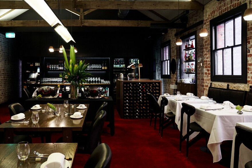 The moody interior of a Chinese restaurant, with exposed brick walls, dark red carpet and tables with white tablecloths on the right and without tablecloths on the left.