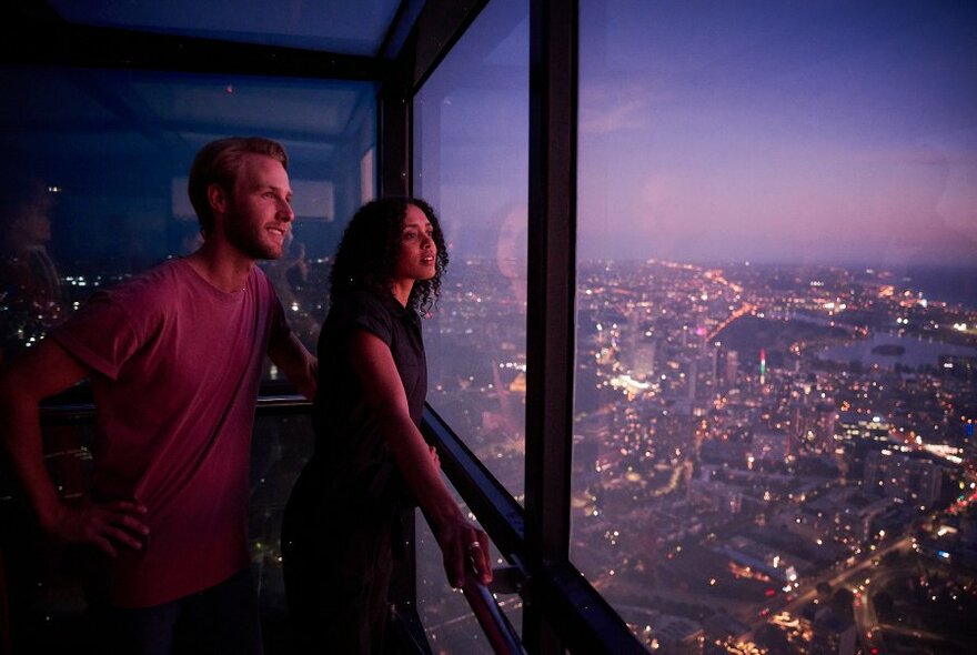 Two people looking out the large glass window at Skydeck high above a twinkling Melbourne city skyline below.