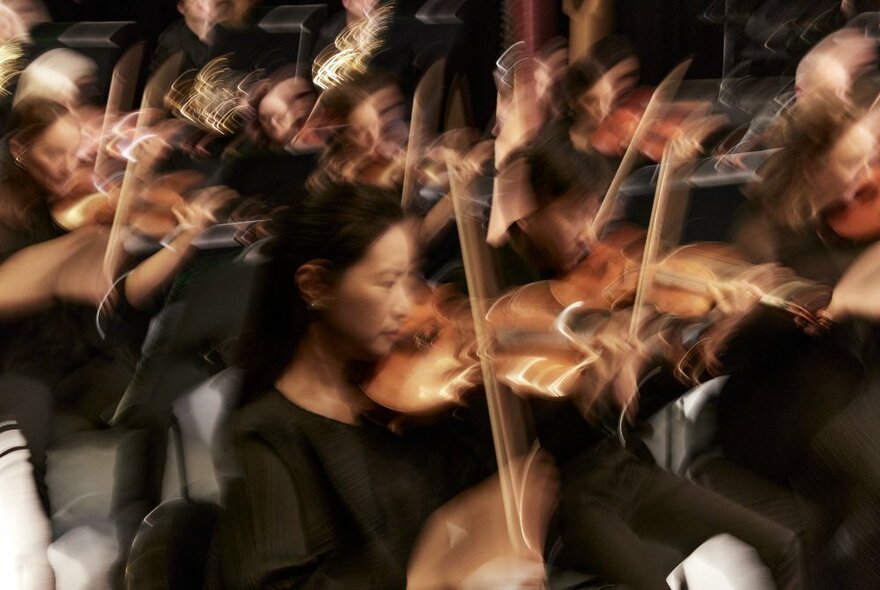 A seated orchestra playing their instruments but blurred to indicate movement and motion of the bow on the strings of the violins and violas.