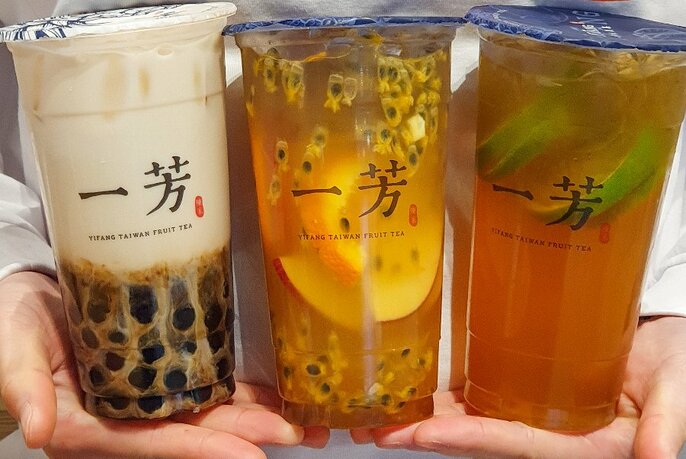 Pair of hands holding up three tall sealed plastic cups of fruit tea. 