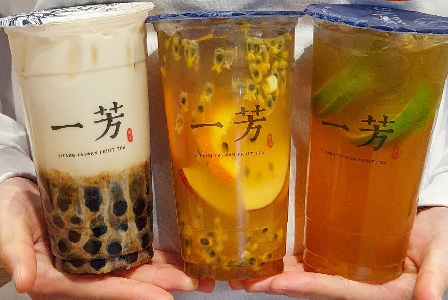 Pair of hands holding up three tall sealed plastic cups of fruit tea. 