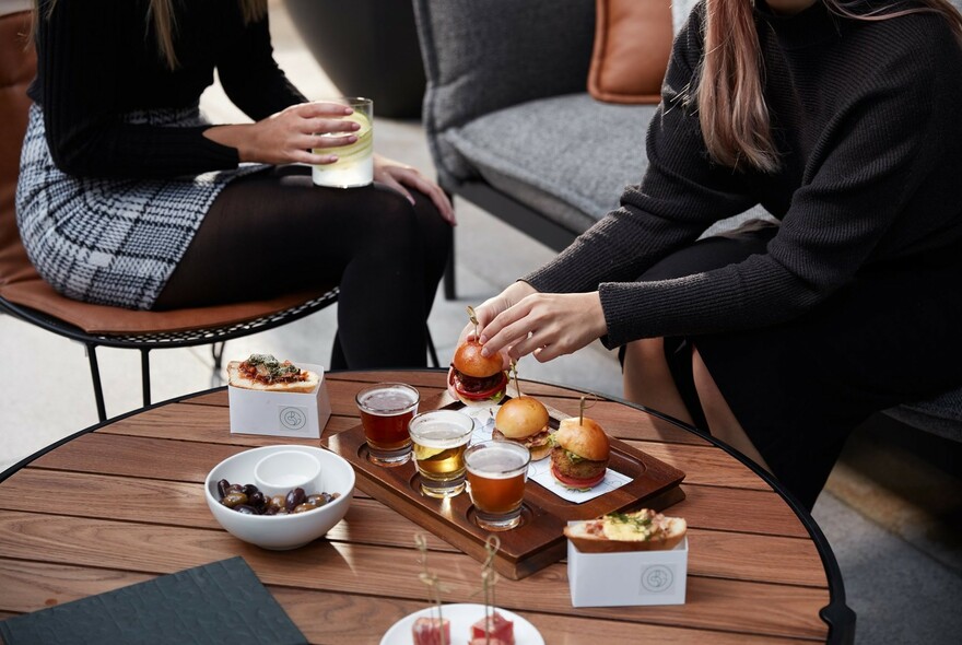 Tray of sliders and drinks on a low table with two patrons partially in view.