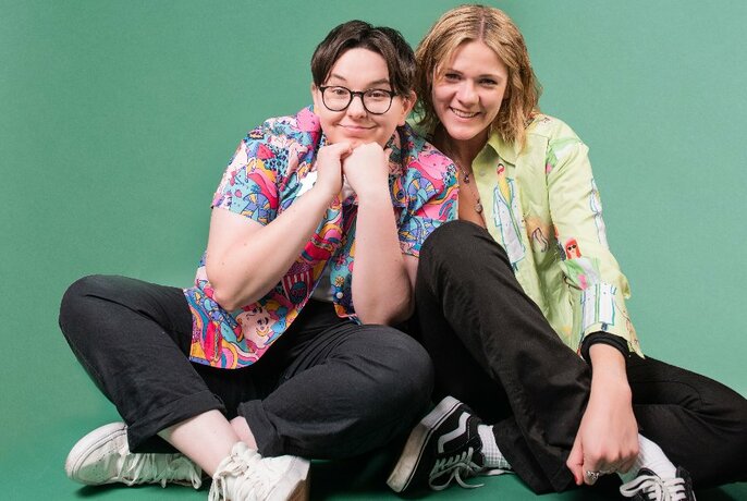 Two comedians smiling and sitting cross-legged on the floor next to each other, against a green background.