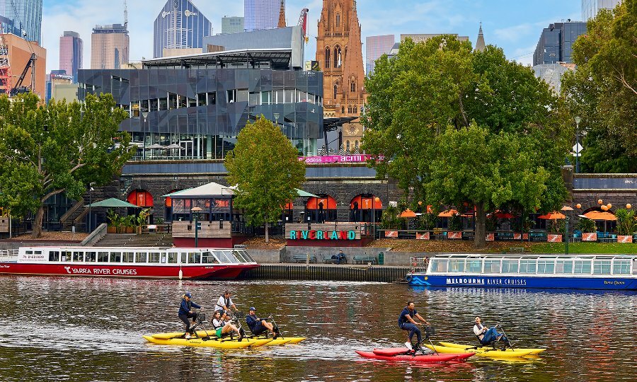 A group of people on colourful water bikes on a city river, with boats moored in the background.