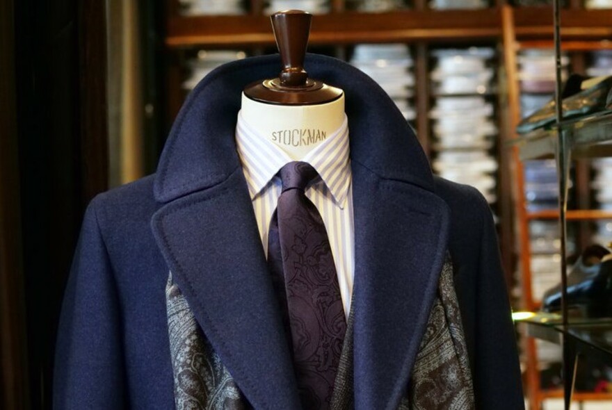 Mannequin wearing blue winter coat, scarf, suit and tie, with rows of shirts in wooden drawers in the background.