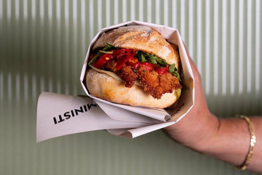 A hand holding a wrapped fried chicken panini in  front of a cream-coloured fluted wall.