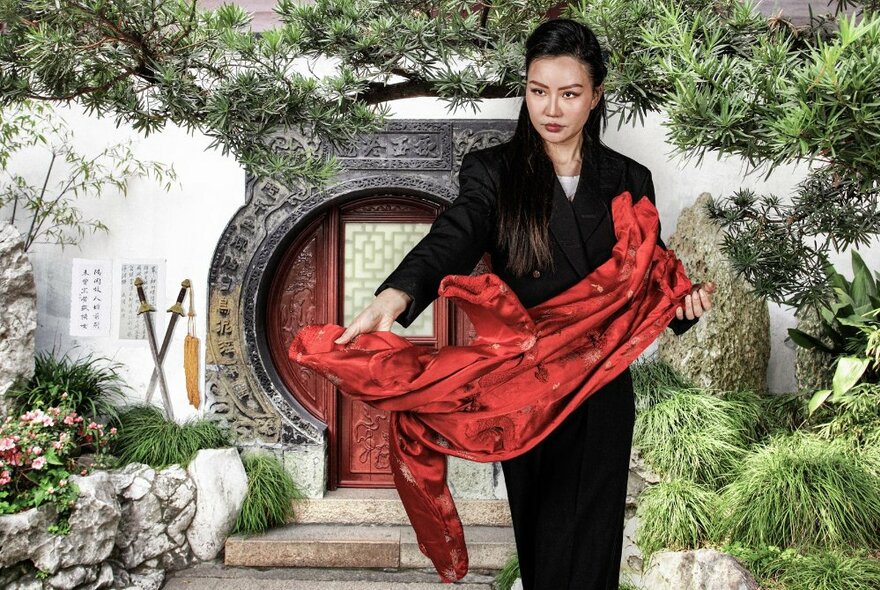 A woman in a traditional Chinese garden, intently staring while holding out a red silk piece of material. 
