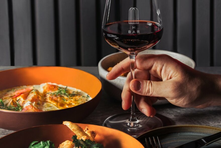 A hand holds the stem of a glass of red wine on a table that holds several orange bowls of food and one white bowl.