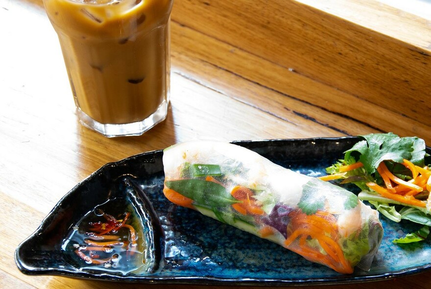 A platter with a vegetable rice-paper roll and iced coffee drink.
