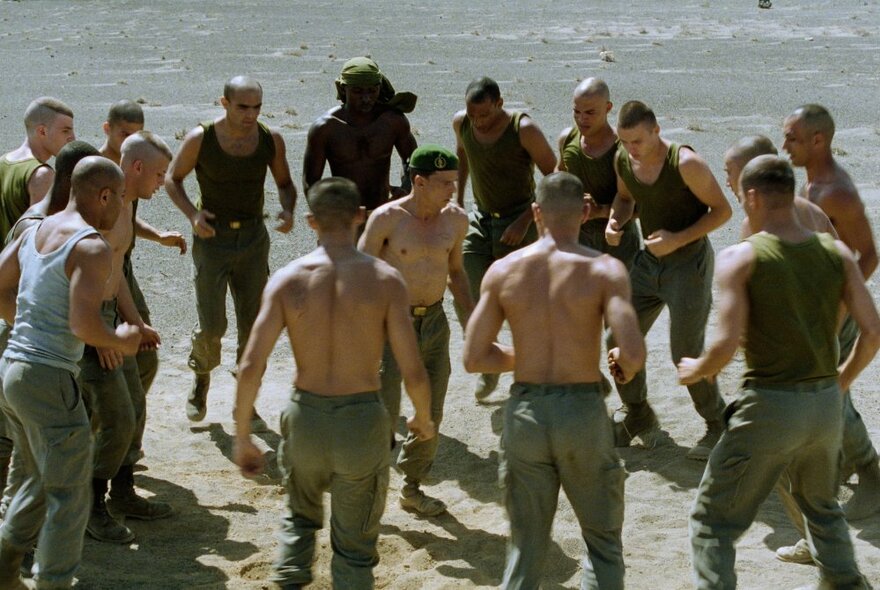 Movie still of rows of bare-chested military members facing each other with arms raised on a stretch of sand.