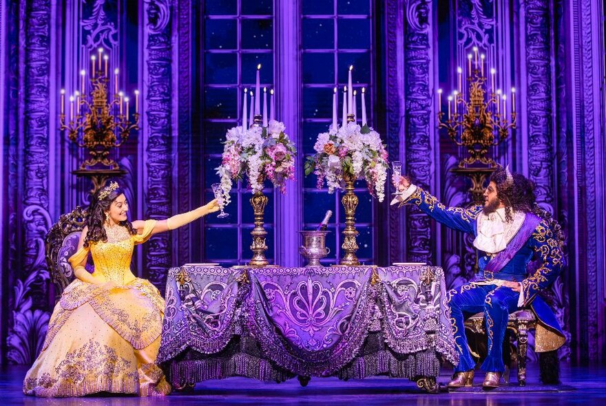 Two cast members from the musical Beauty and the Beast - the beast and the beauty - on stage, sitting at opposite sides of a dining table and raising their glasses in a toast.