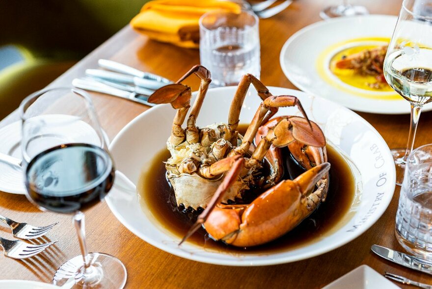 Plate of whole mud crab in a sauce, on a table with glasses of wine, napkin and cutlery.