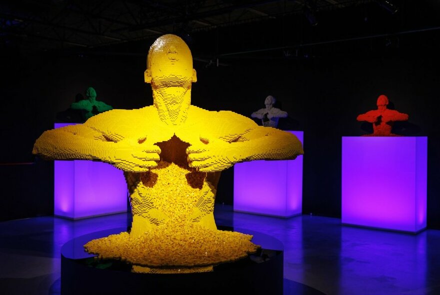 Sculpture of a torso pulling its chest open, crafted from many yellow lego bricks, on display in a large exhibition space.