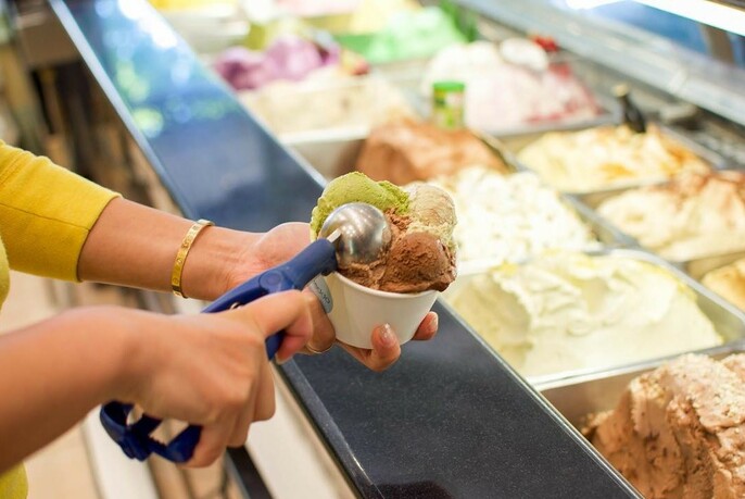 A person scooping ice cream into a tub with many different flavours in the background. 