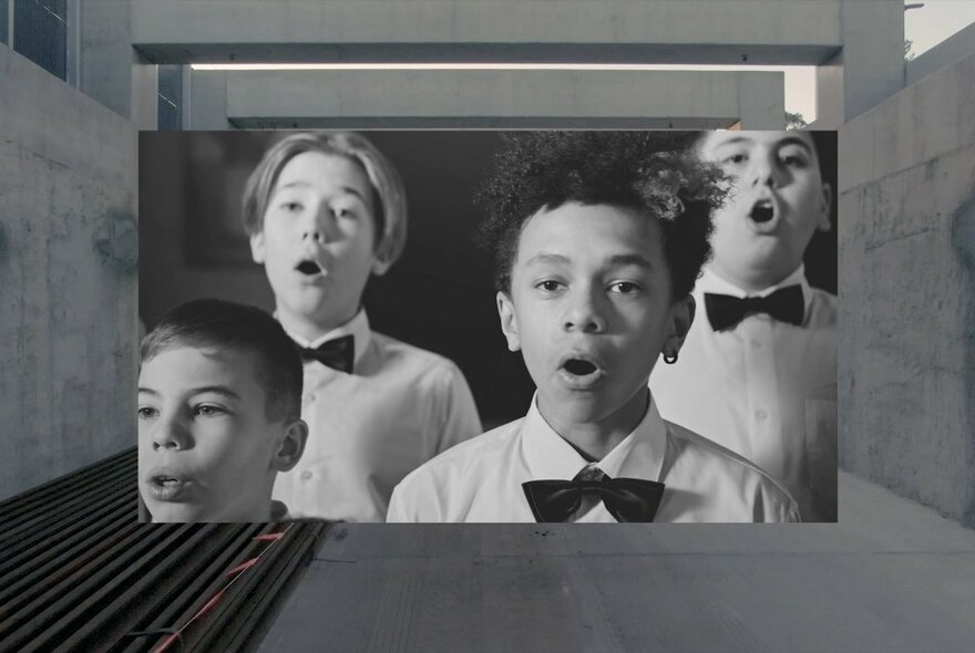 A black and white picture of four choir boys in white shirts and black bow-ties, singing, placed on top of a background image of a concrete tunnel.