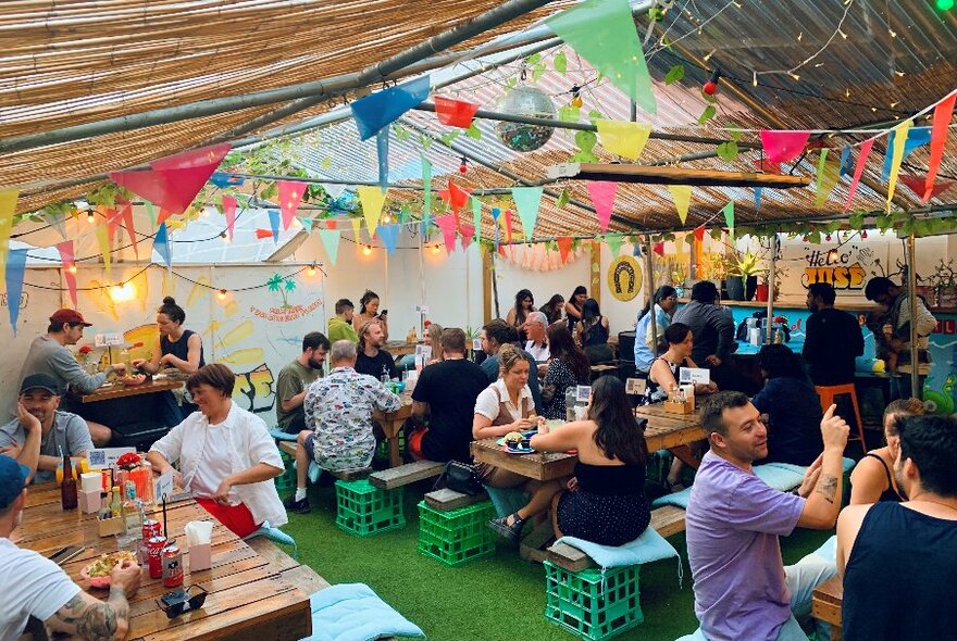 People seated on milk-crate benches at outdoor tables under bunting and a fabric roof in a beer garden atmosphere.