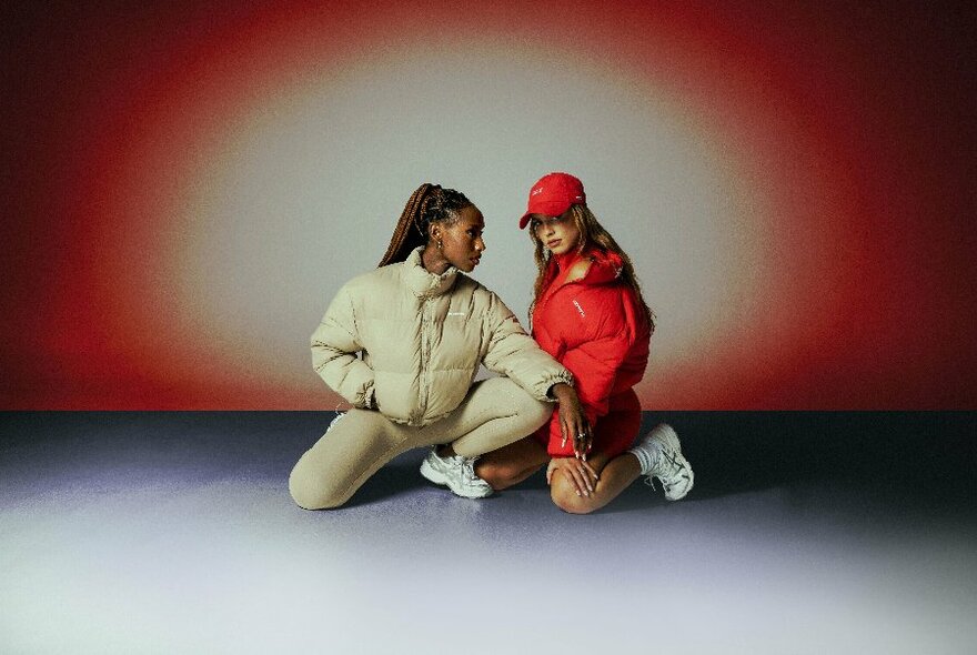 Two women wearing sports leggings and puffy jackets, kneeling on the ground.