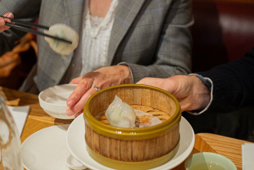 Hands holding dumplings with chopsticks next to a bamboo steamer in a restaurant.