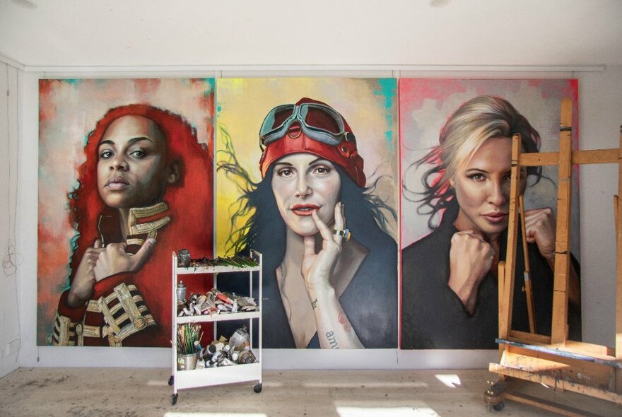 Three large portraits of women, triptych style, on the rear of a studio wall, an easel and a trolley of painting utensils also in the room.