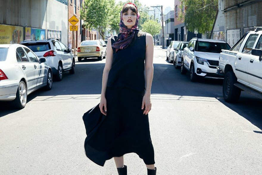 A model standing on a road wearing a black flowy midi dress, headscarf and boots.