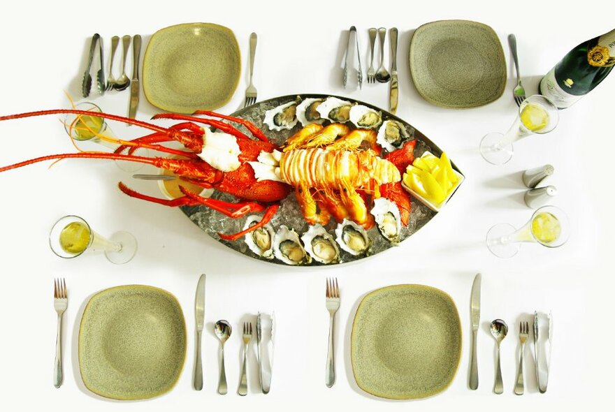 A dining setting on a white tablecloth with olive green plates and cutlery, an elaborate lobster dish with oysters in the centre of the table with glasses of white wine.