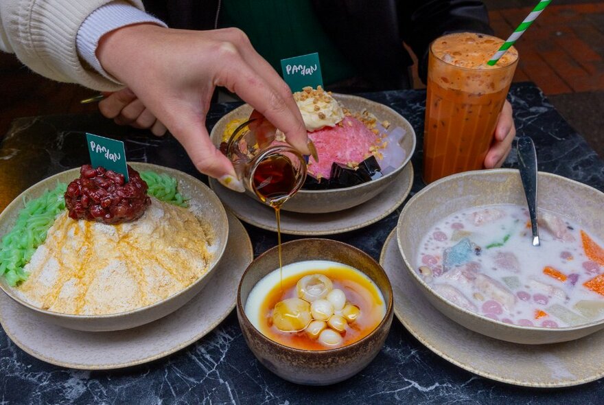 A hand pouring sweet syrup from a small glass onto a Malaysian dessert served in a bowl, other colorful desserts in large bowls on the table, as well as an orange drink in a tall glass.