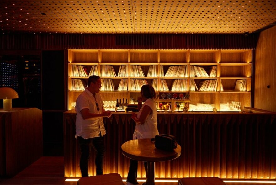 A dimly lit bar with two people talking, a wall of records on shelves behind the bar where they are standing.